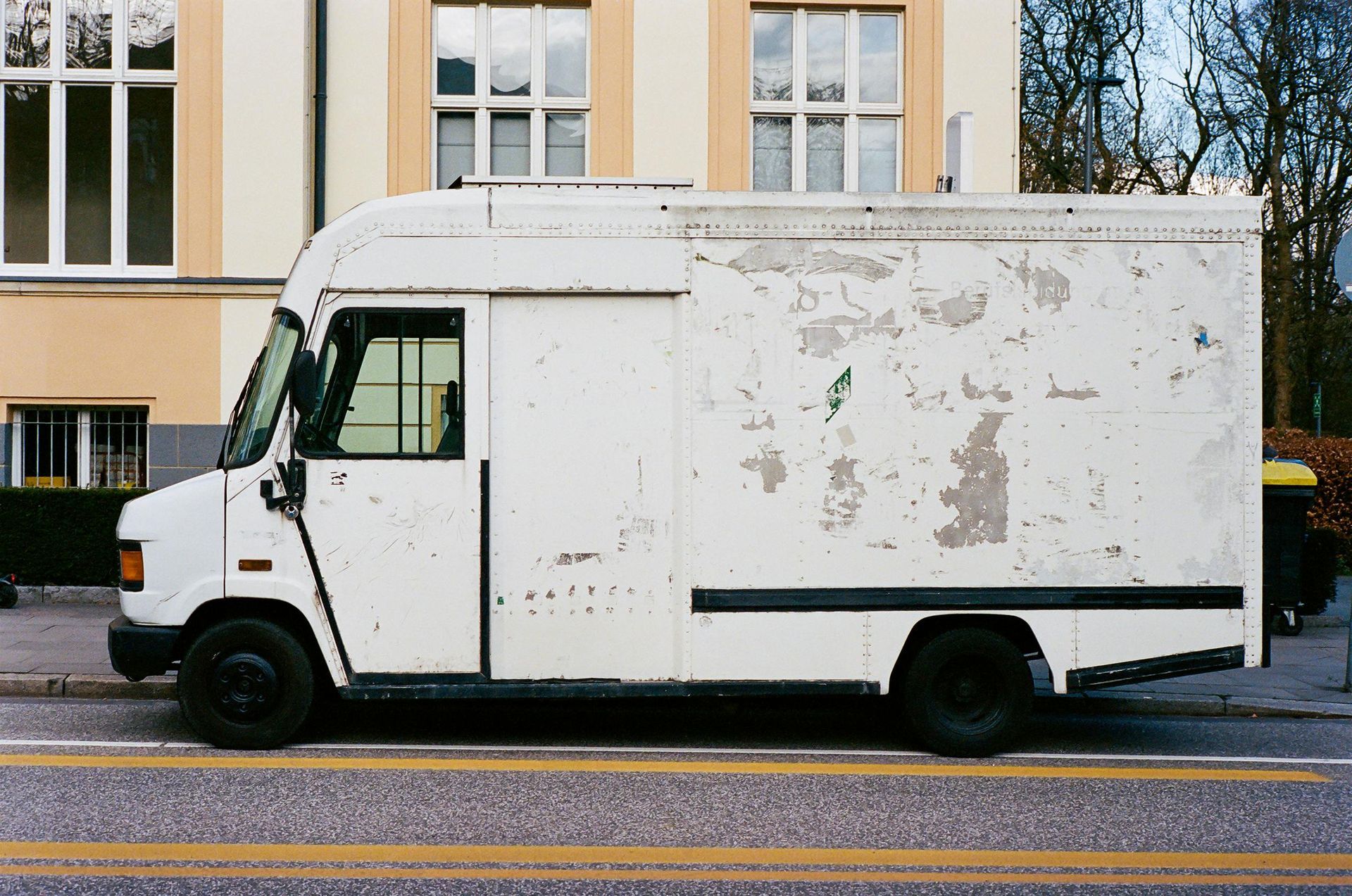 A white box truck with chipped paint is parked on a paved street in front of a building with large, multi-pane windows.