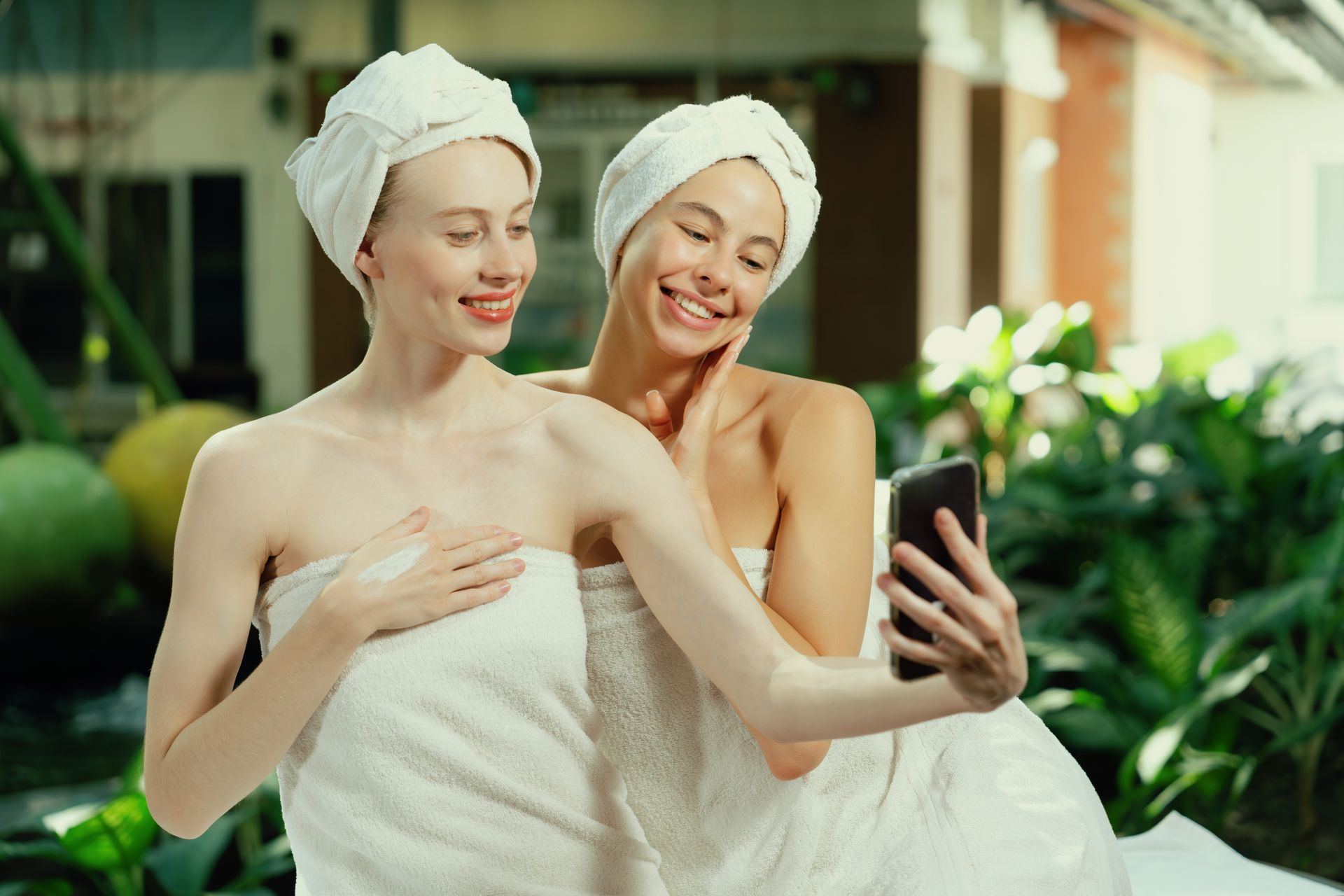 Two women wrapped in towels take a selfie, smiling, outside with green foliage.