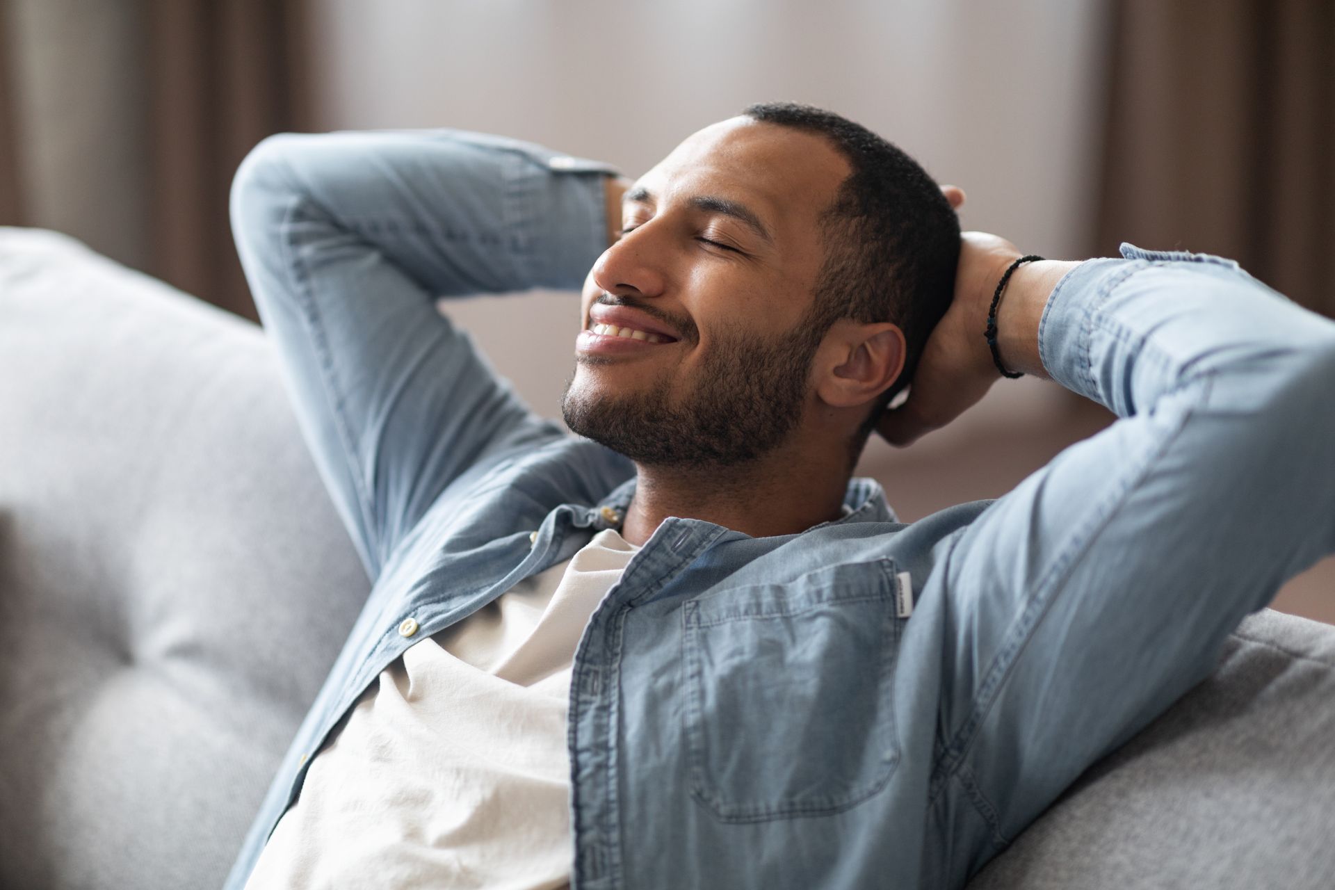 A man is sitting on a couch with his hands behind his head and smiling.