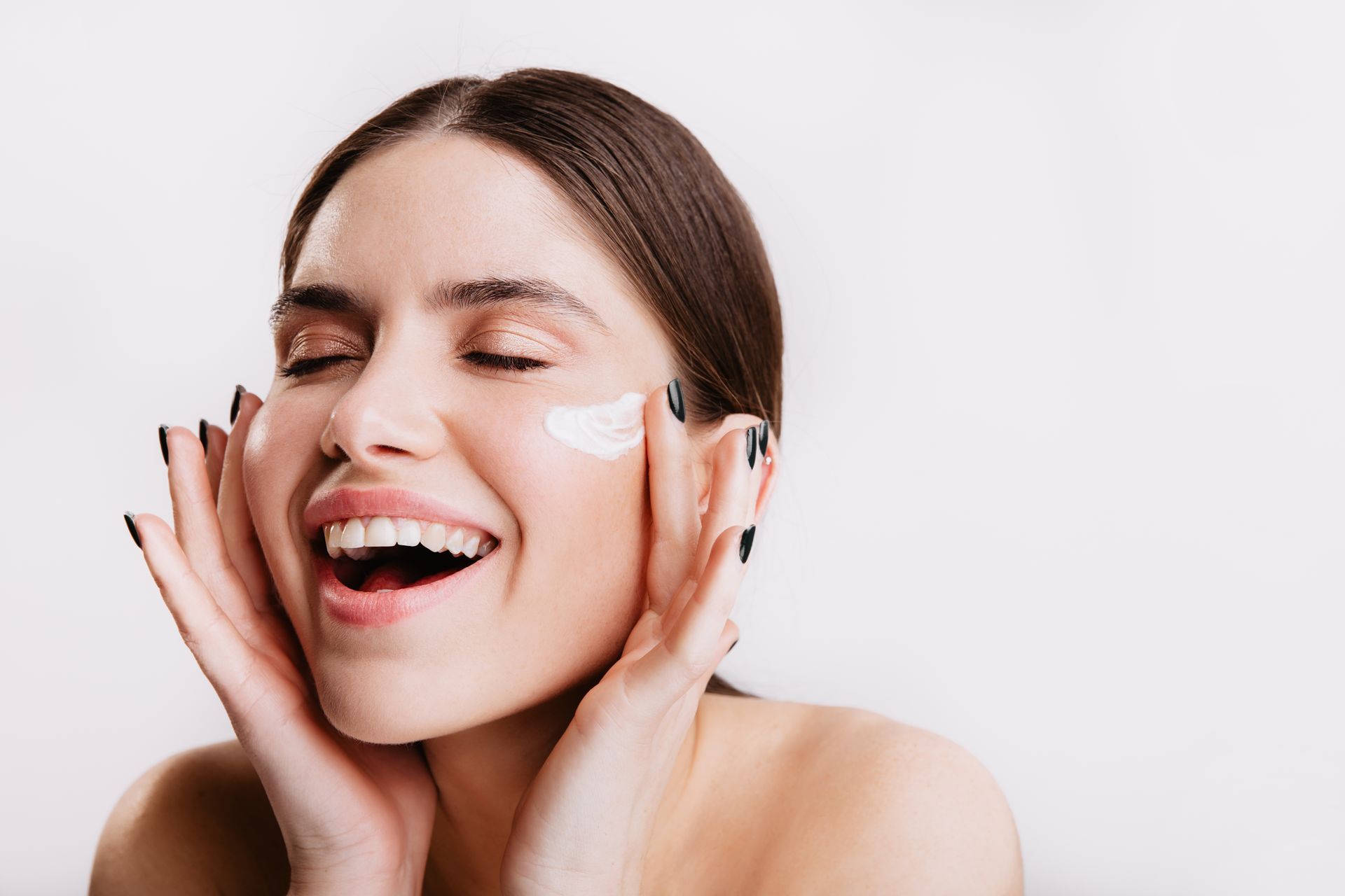 A woman is smiling while applying cream to her face.