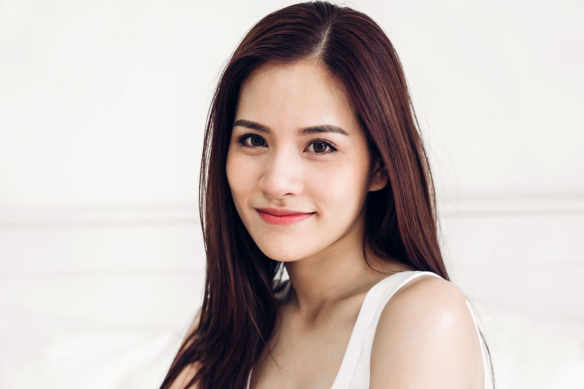 A woman with long brown hair is wearing a white tank top and earrings.