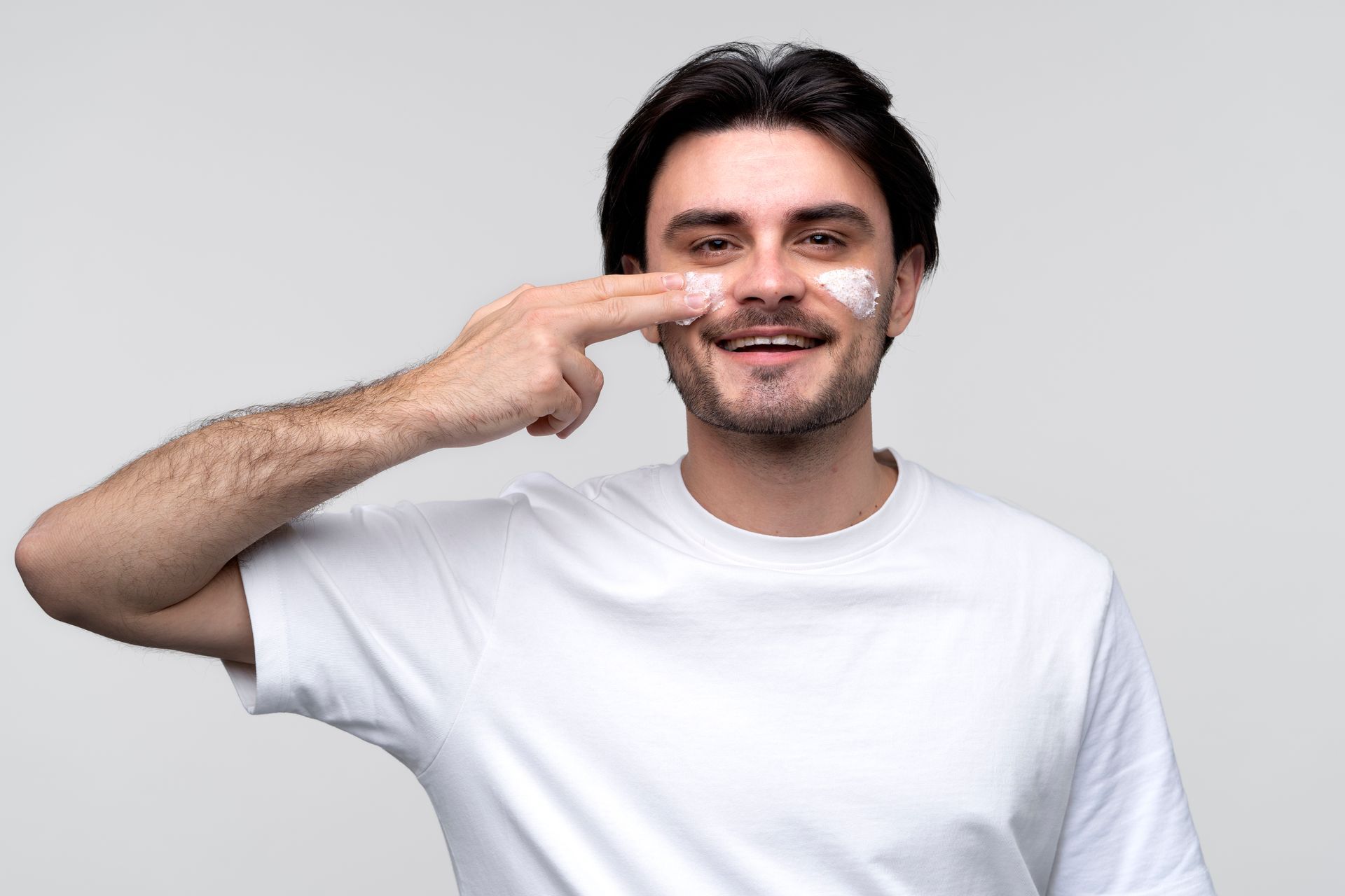 A man in a white t-shirt is applying cream to his face.