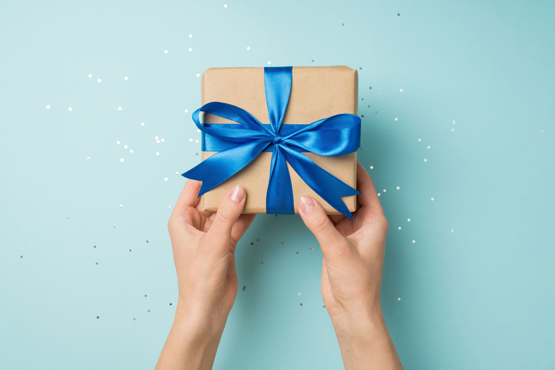 Hands holding a wrapped gift with a blue bow on a light blue background.