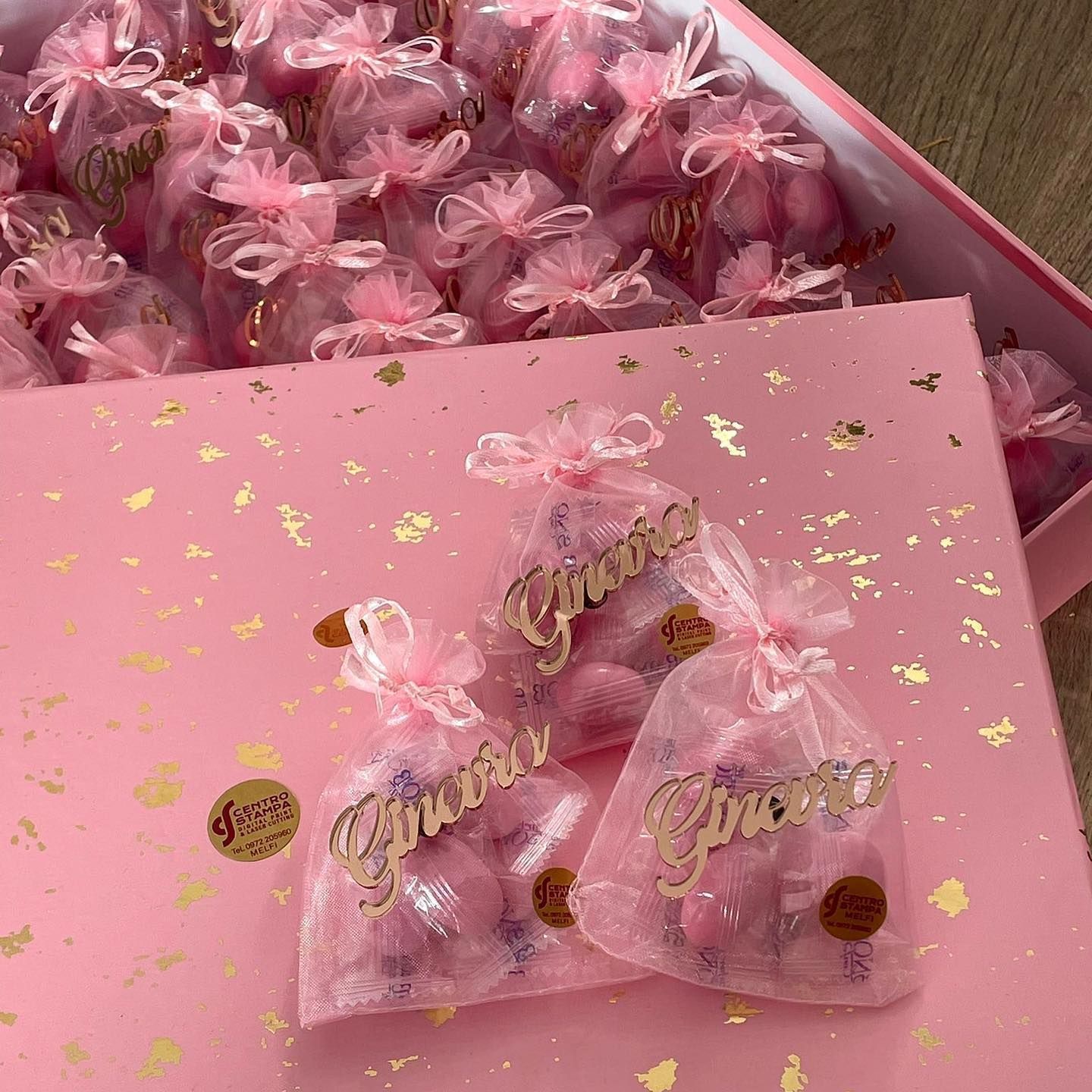 Sacchetti di organza rosa pieni di caramelle, legati con nastri, in una scatola rosa con dettagli dorati.