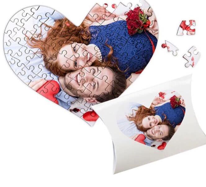 Puzzle a forma di cuore con la foto di una coppia, parzialmente assemblato, accanto a una scatola regalo coordinata.