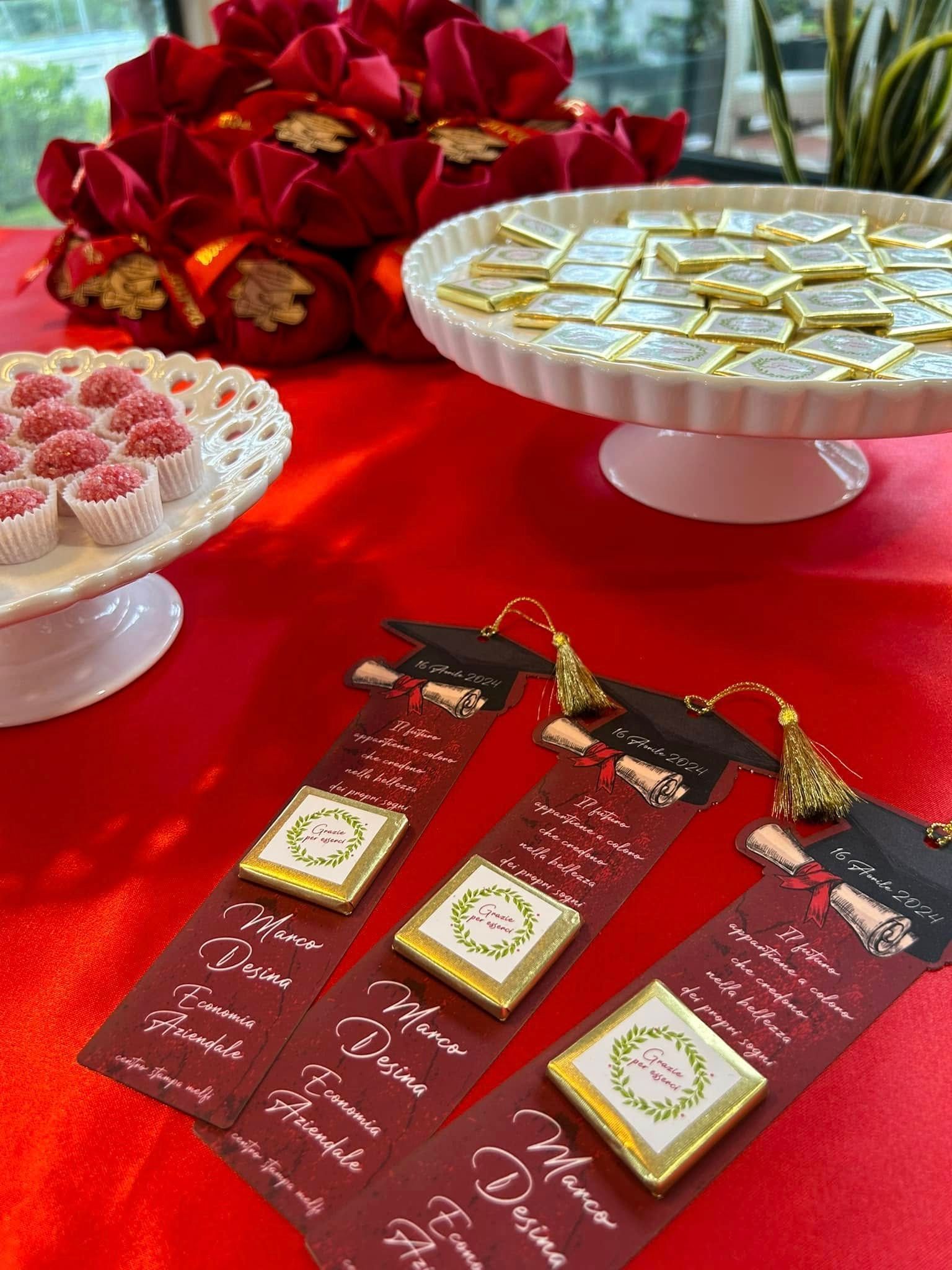 Dolcetti per la festa di laurea: tavolo rosso con dolci, bomboniere, barrette di cioccolato e composizioni floreali.