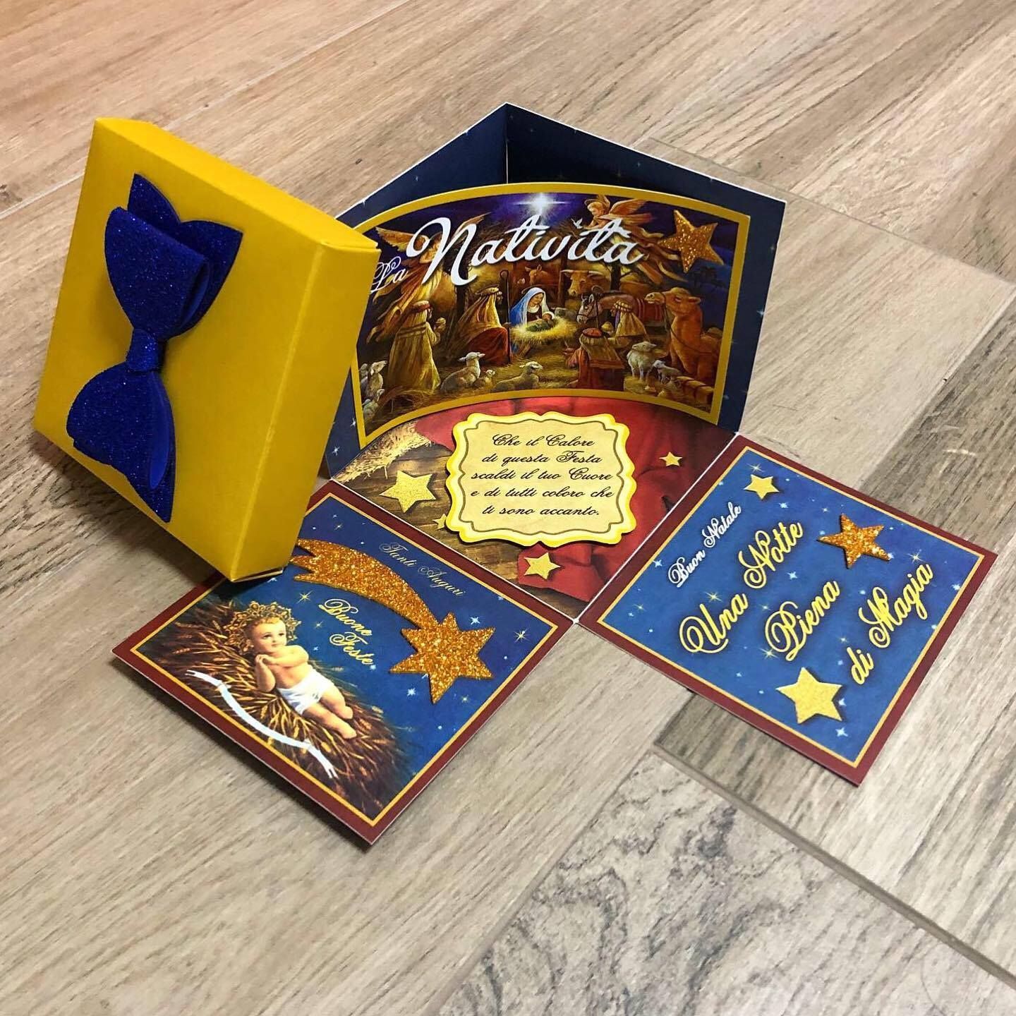 Biglietti di auguri di Natale e una scatola gialla con un fiocco blu; i biglietti raffigurano un presepe, un bambino e auguri in italiano.