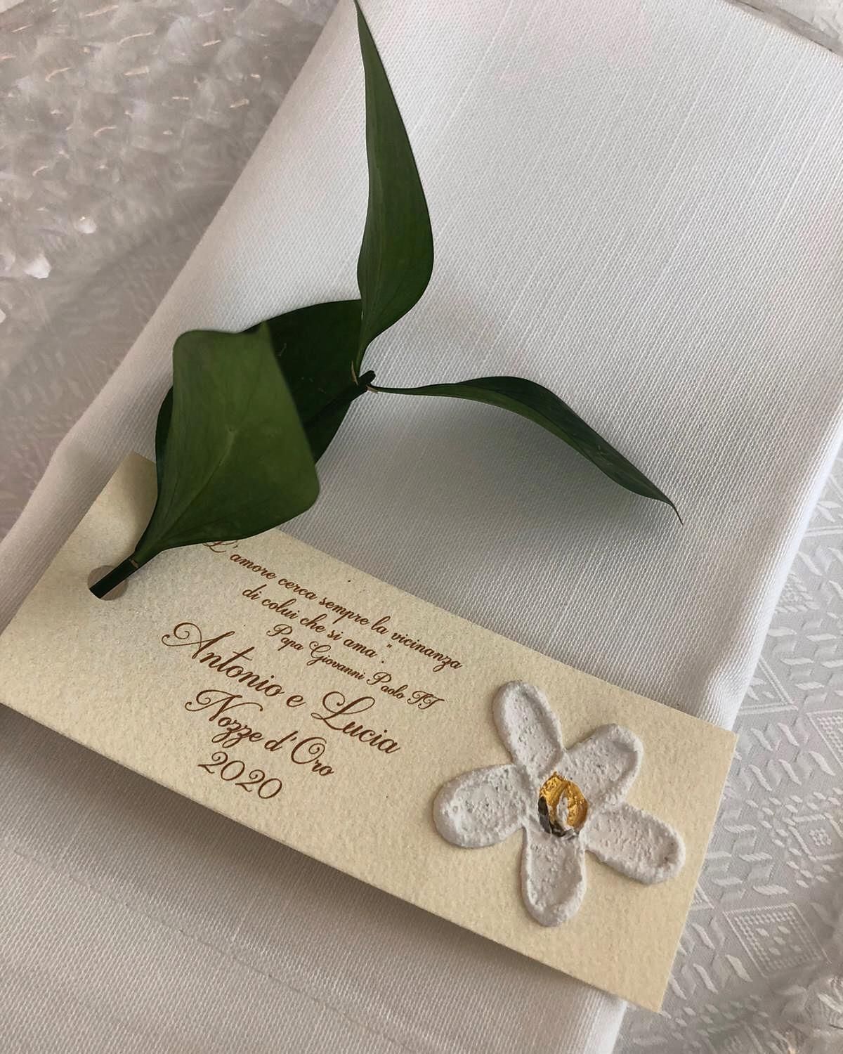 Tovagliolo bianco con un biglietto e del verde. Il biglietto ha un fiore bianco testurizzato e i nomi: Antonio e Lucia, 2020.