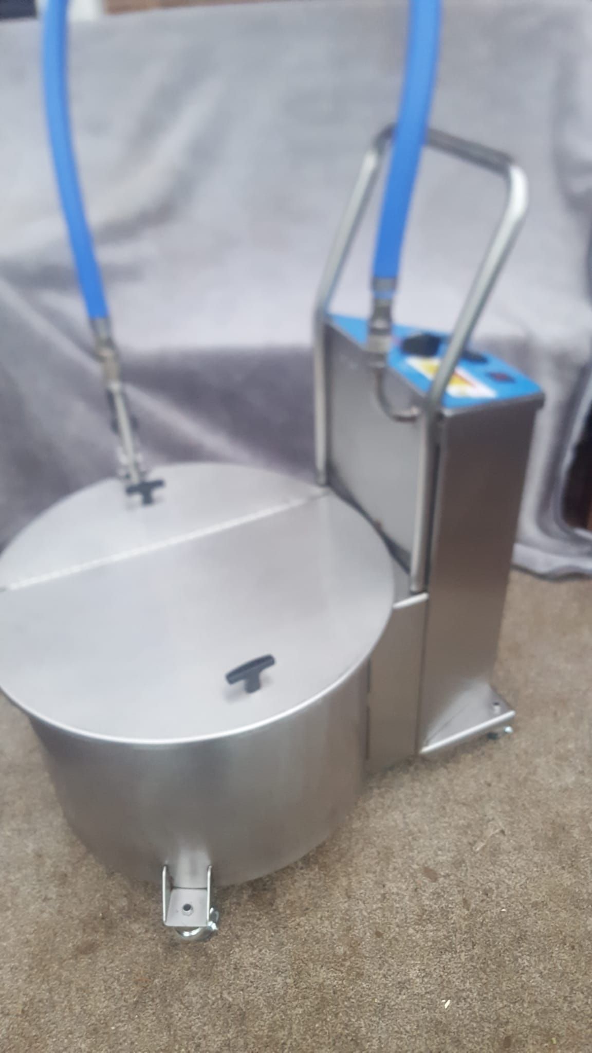 A stainless steel bucket with two blue hoses attached to it is sitting on a carpet.