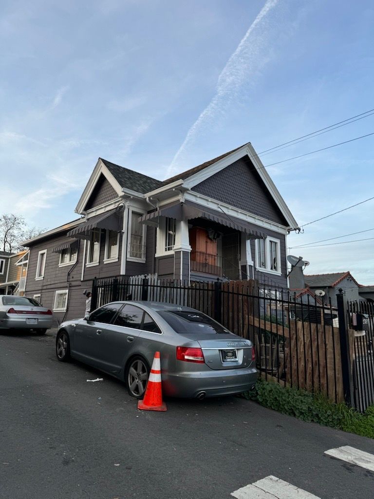 Gray house with silver car parked on an inclined street; orange traffic cone sits nearby. Gray house with silver car parked on an inclined street; orange traffic cone sits nearby.