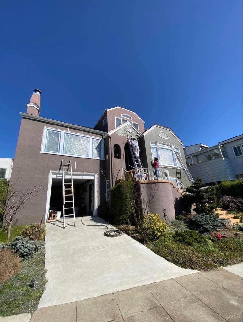 House exterior being painted, with ladders and painters visible. Dusty rose stucco with blue sky. House exterior being painted, with ladders and painters visible. Dusty rose stucco with blue sky.