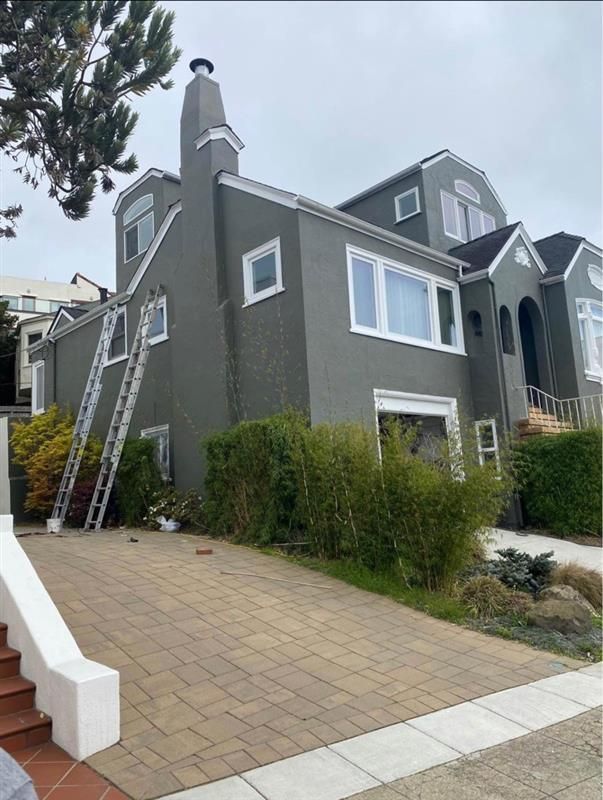Gray house with a ladder leaning against it, driveway in front, and bushes.