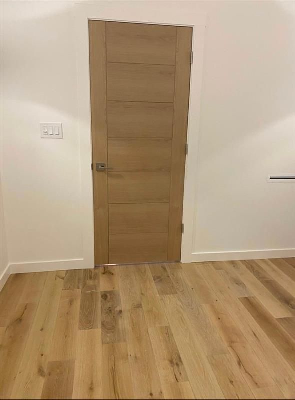 Wooden door in a room with light wood flooring and white walls. Light switch on the left wall.