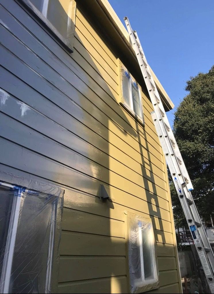 Side of a house being painted, with a tall ladder extending to the roof. Windows are taped, exterior is yellow-green. Side of a house being painted, with a tall ladder extending to the roof. Windows are taped, exterior is yellow-green.