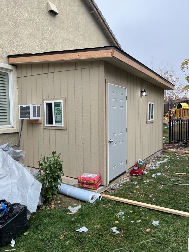 Tan shed with a white door, windows, and an AC unit attached to a beige house, surrounded by grass. Tan shed with a white door, windows, and an AC unit attached to a beige house, surrounded by grass.