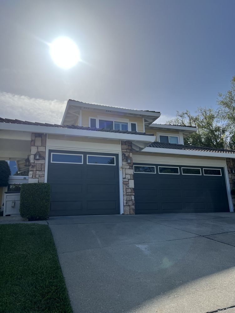 Two-car garage with dark gray doors and windows. Beige house with stone accents on sunny day. Two-car garage with dark gray doors and windows. Beige house with stone accents on sunny day.