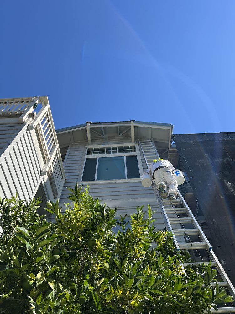 Person in protective suit on ladder, working on a house exterior under a blue sky. Person in protective suit on ladder, working on a house exterior under a blue sky.