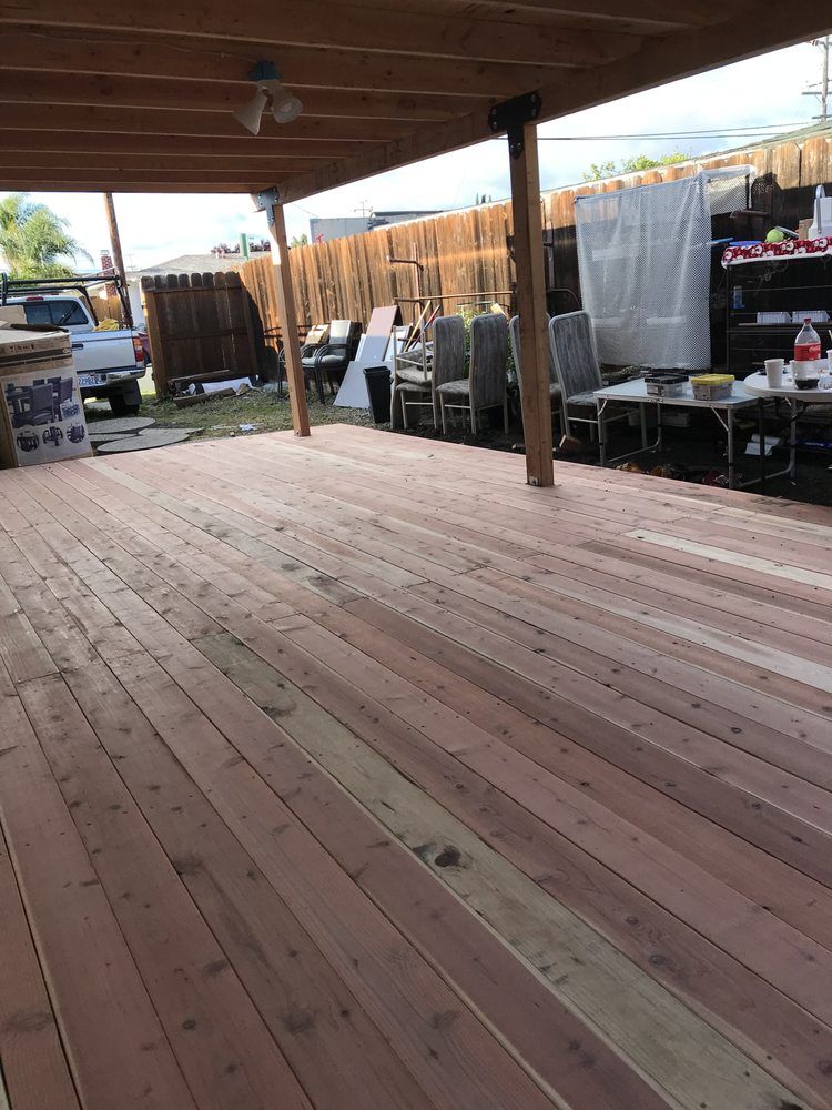 Wooden deck under a wooden pergola, partially shaded. Various items and fence in the background.