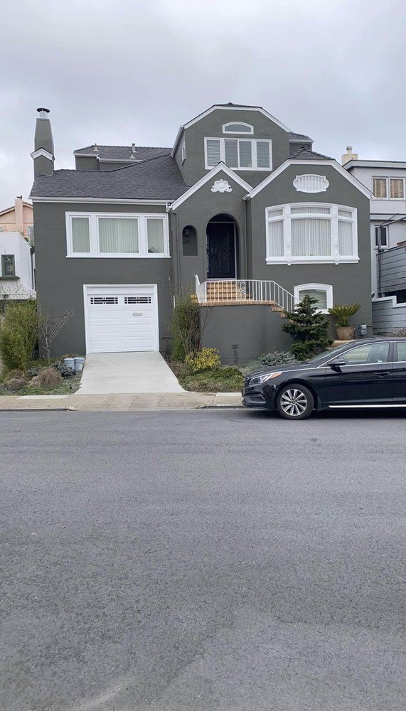 Gray house with white trim, garage door, and a black car parked on the street. Overcast sky.