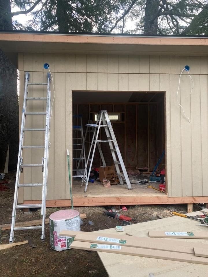 Construction of a shed with an open doorway, ladders, and tools scattered around. Light brown siding, neutral tones.