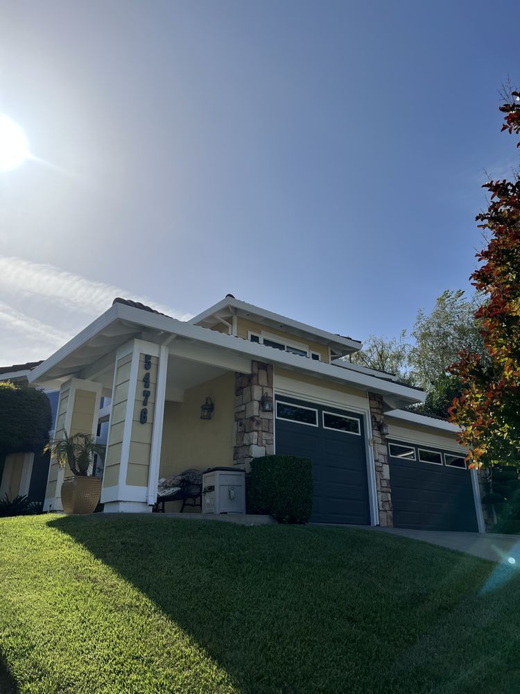 Yellow house with attached garage, stone accents, dark garage doors, and lush green lawn under a bright blue sky.