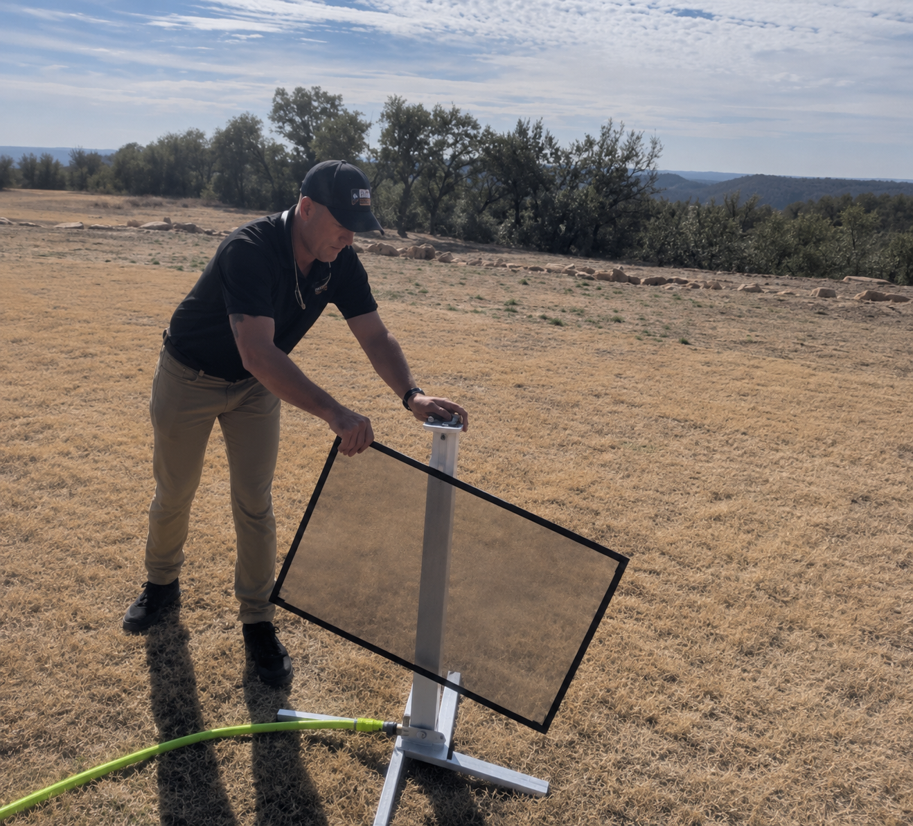 Person adjusting a large tilted solar panel outdoors on dry grass, with trees and open sky in the background