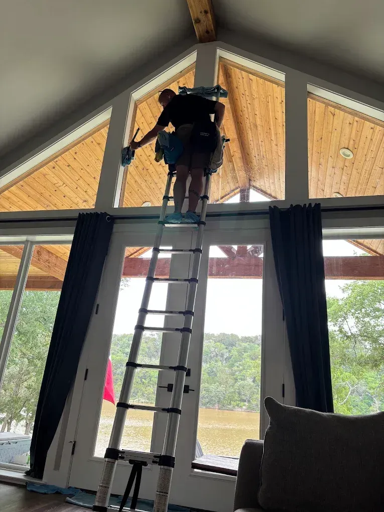 Person on a ladder adjusting ceiling wiring in a room with large windows and wood-paneled walls