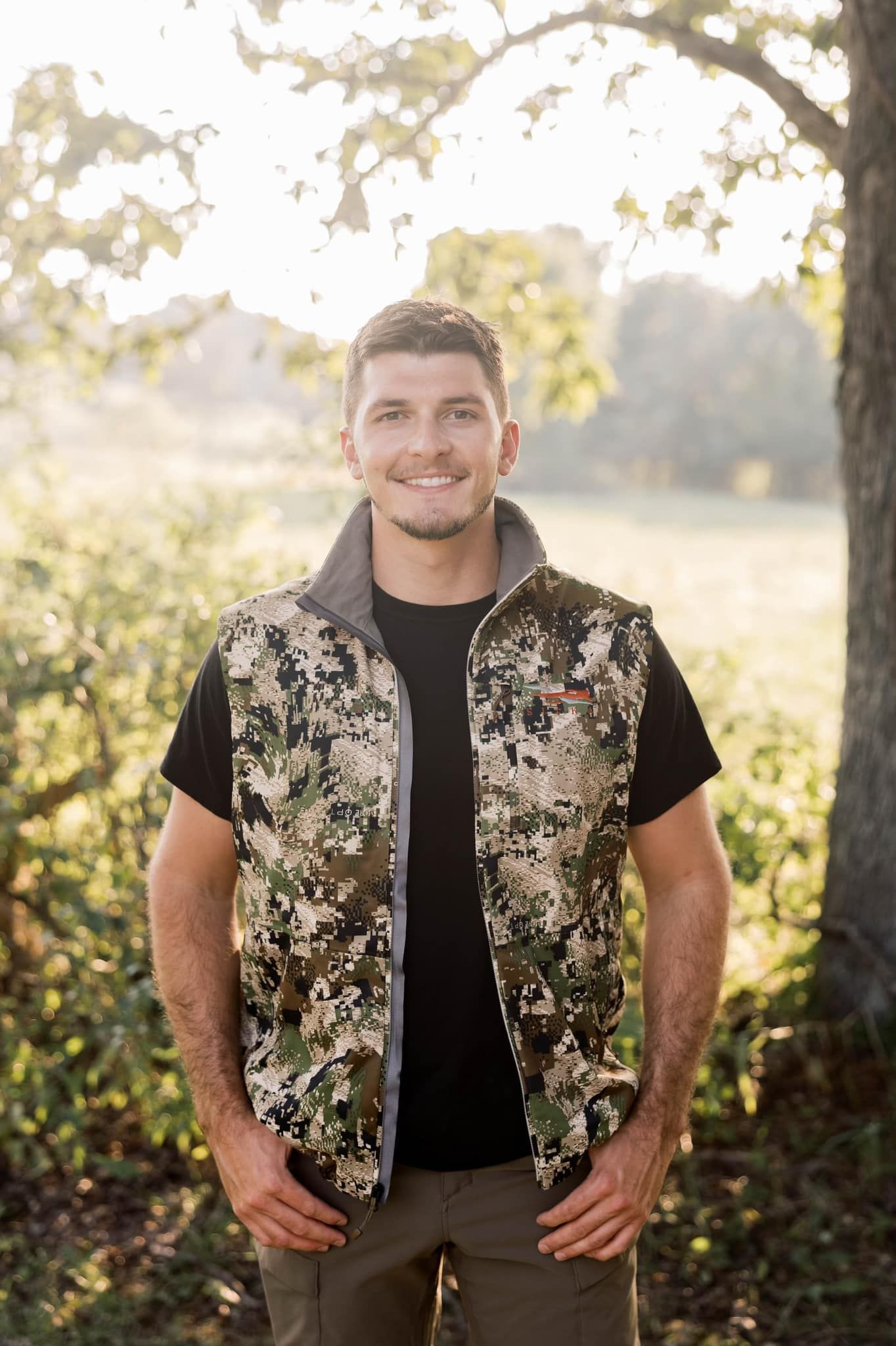 Two men pose with a harvested deer. They're sitting in tall grass, one with a rifle, both smiling, under a dark sky.