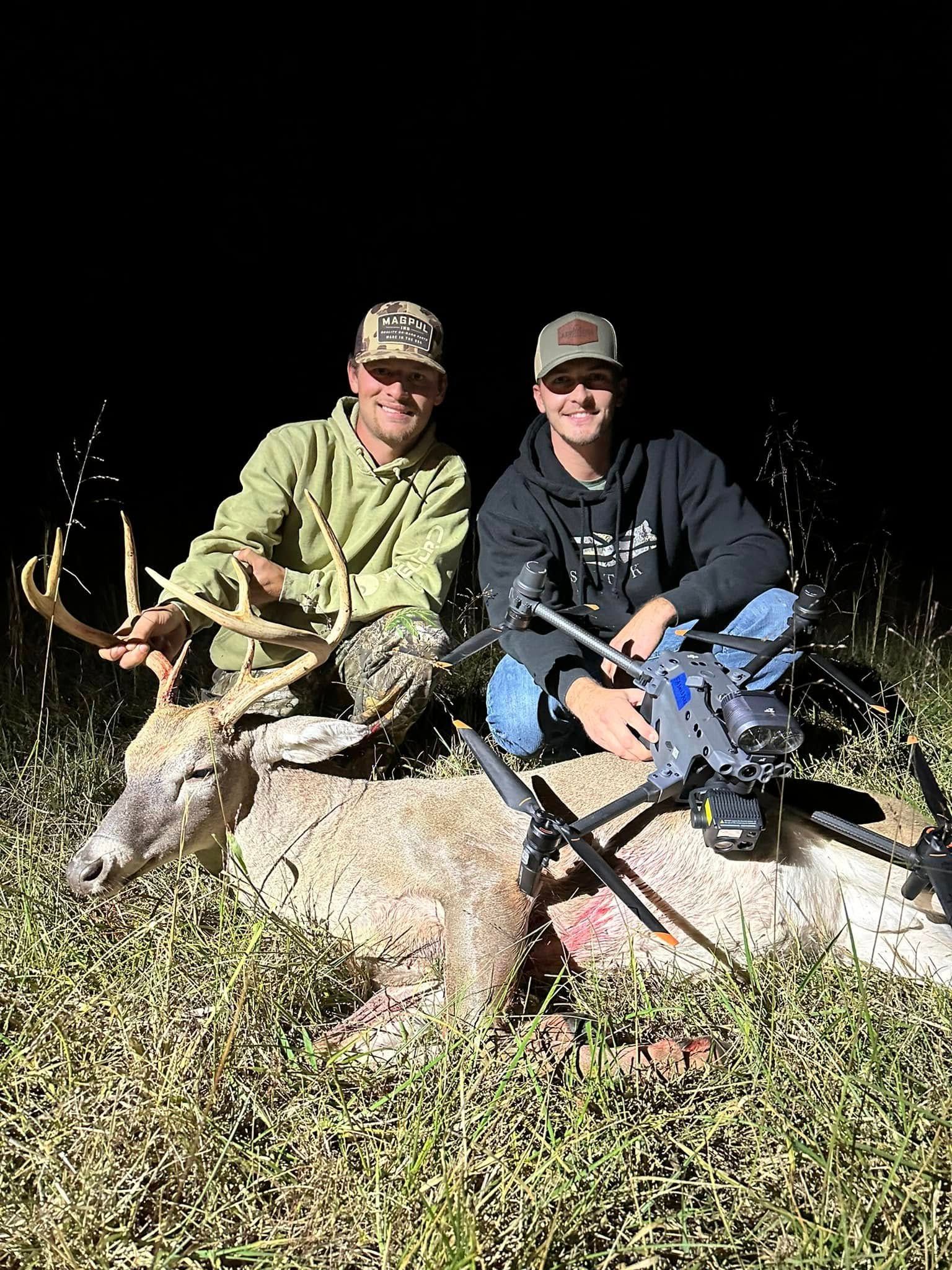 Two men pose with a harvested deer. They're sitting in tall grass, one with a rifle, both smiling, under a dark sky.