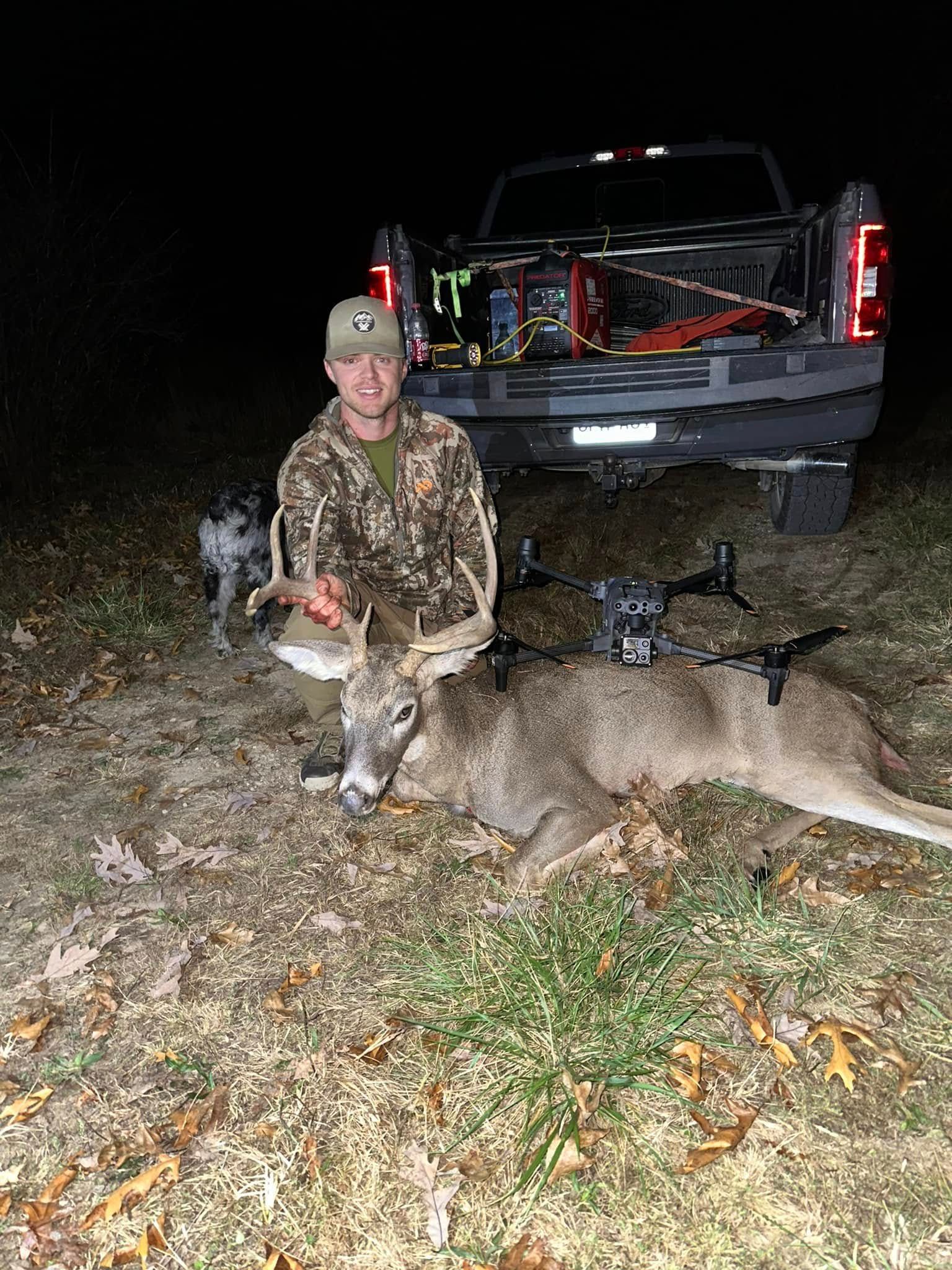 Man in camo with a harvested deer and bow in front of a truck at night.