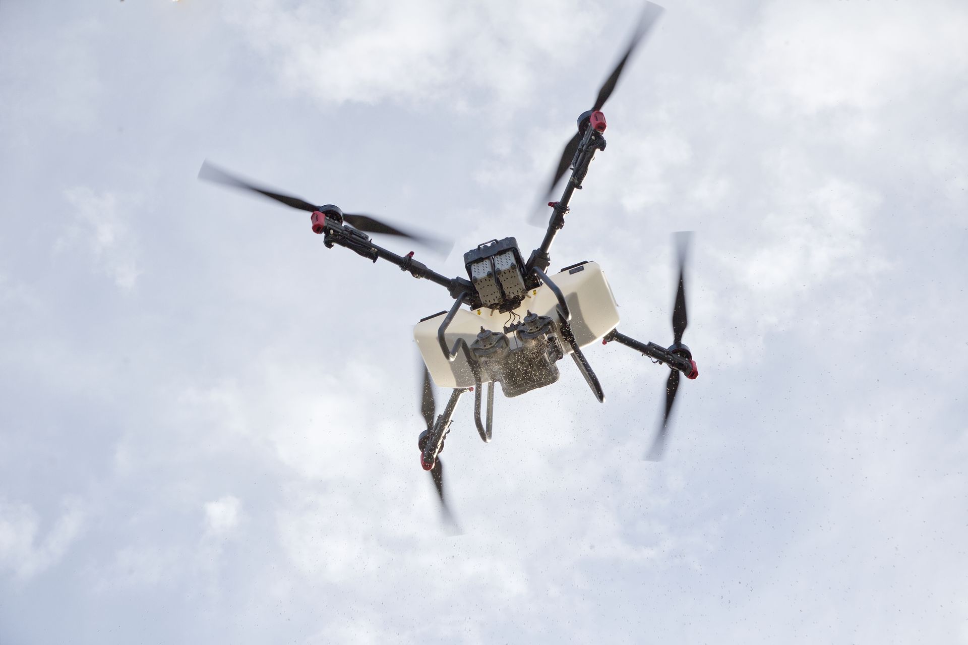 Drone spraying a field, propellers spinning, against a cloudy sky.