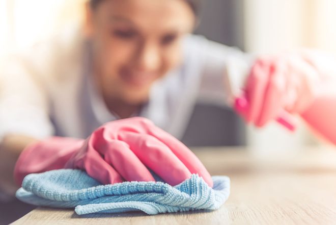 Woman in protective gloves is smiling and wiping dust using a spray and a duster while cleaning her house