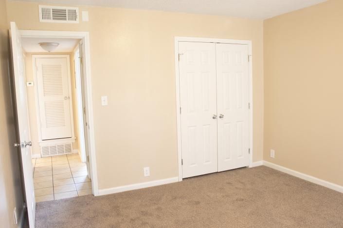 Empty beige bedroom with carpet, white double closet doors, and hallway visible through an open doorway