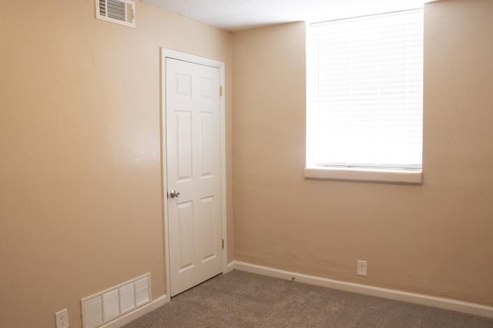 Empty beige bedroom with white door, window blinds, and carpeted floor