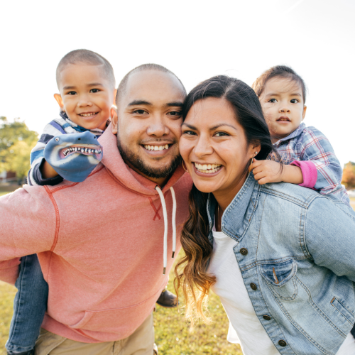 Smiling family of four outdoors, with two children on parents’ backs in bright sunlight