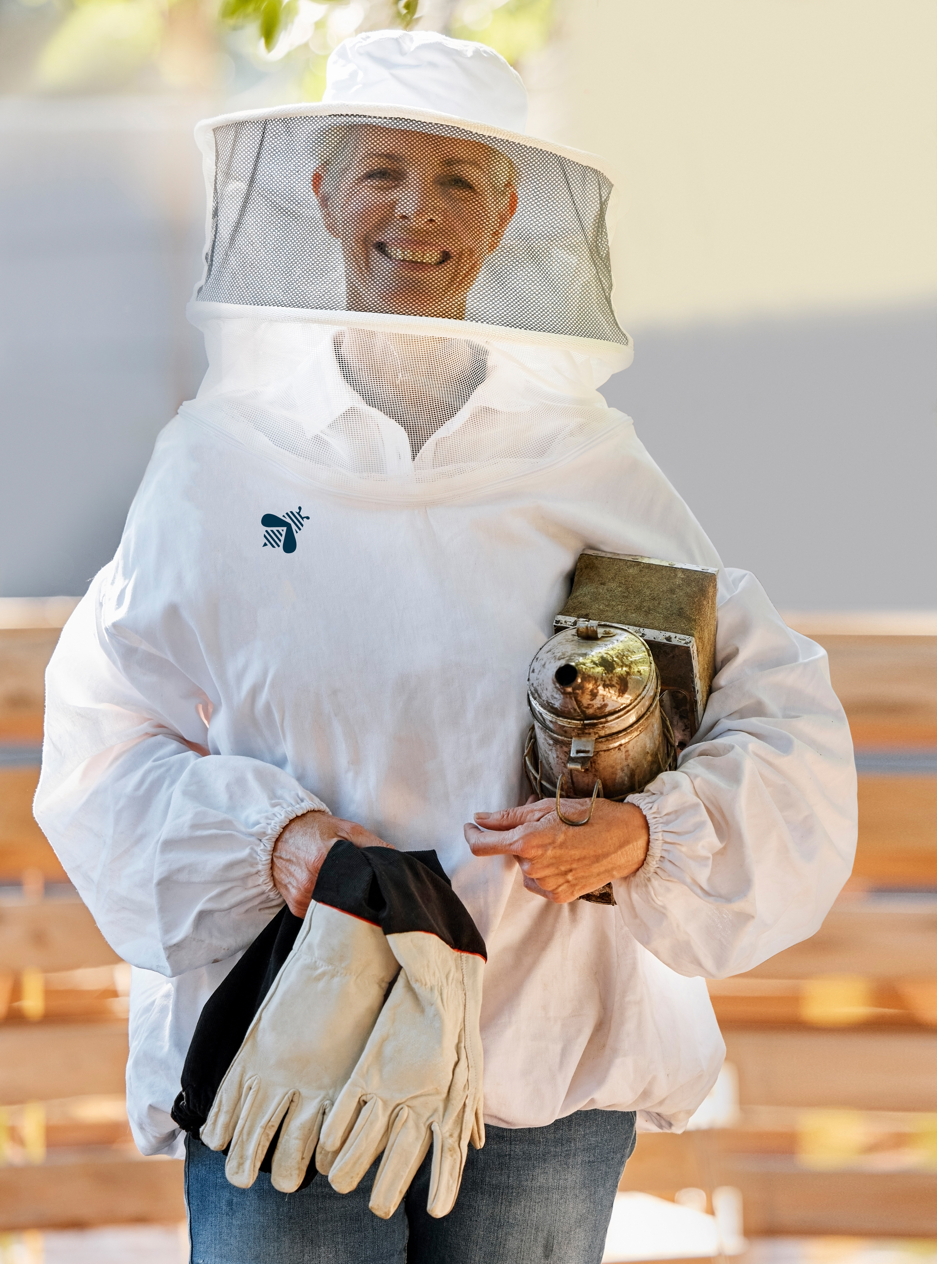 Person in a white beekeeping suit holds gloves and a smoker; outdoors.