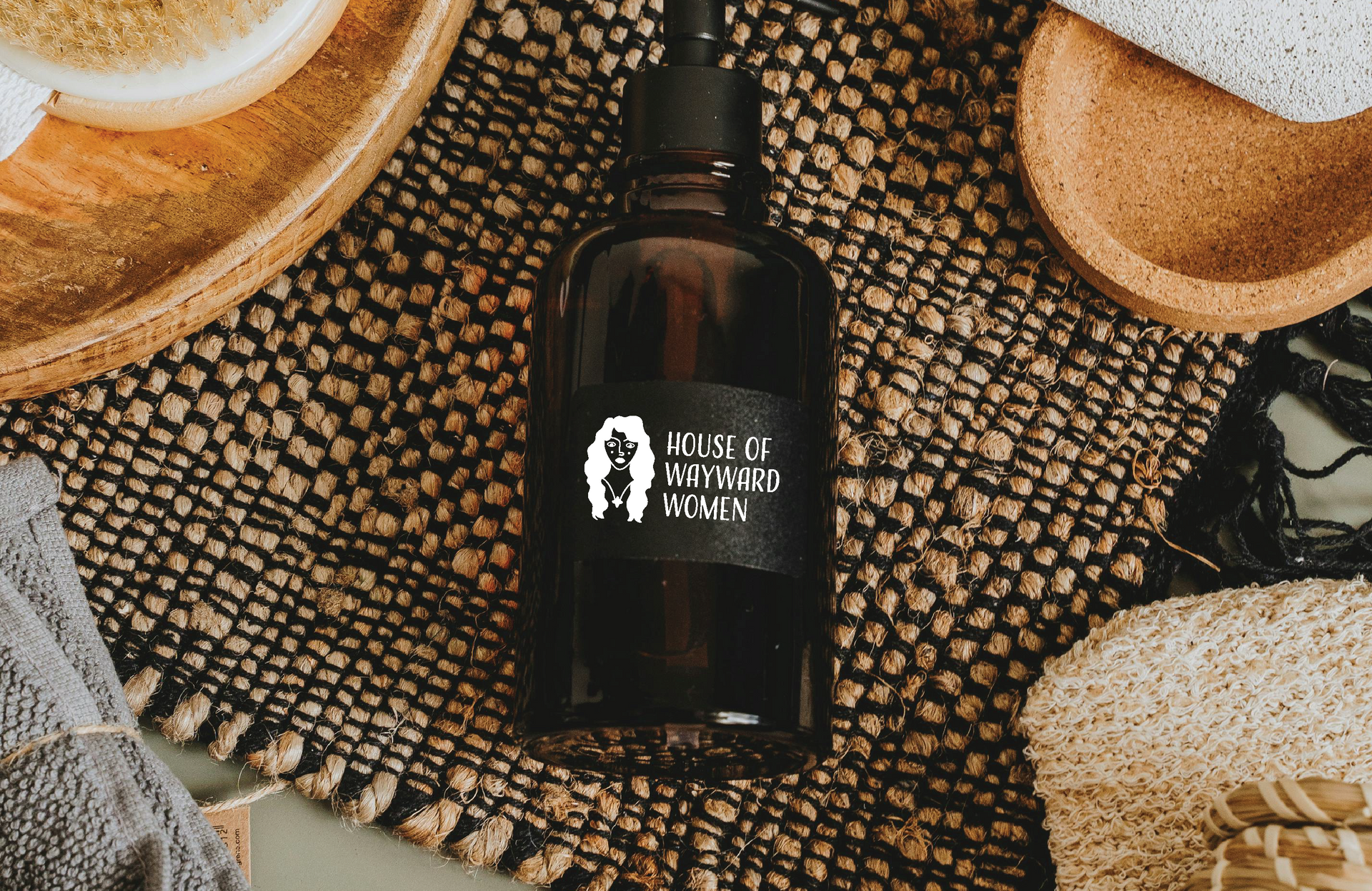 Dark glass bottle with a white House of Wayward Women label, on a woven mat with soap and scrub brushes.