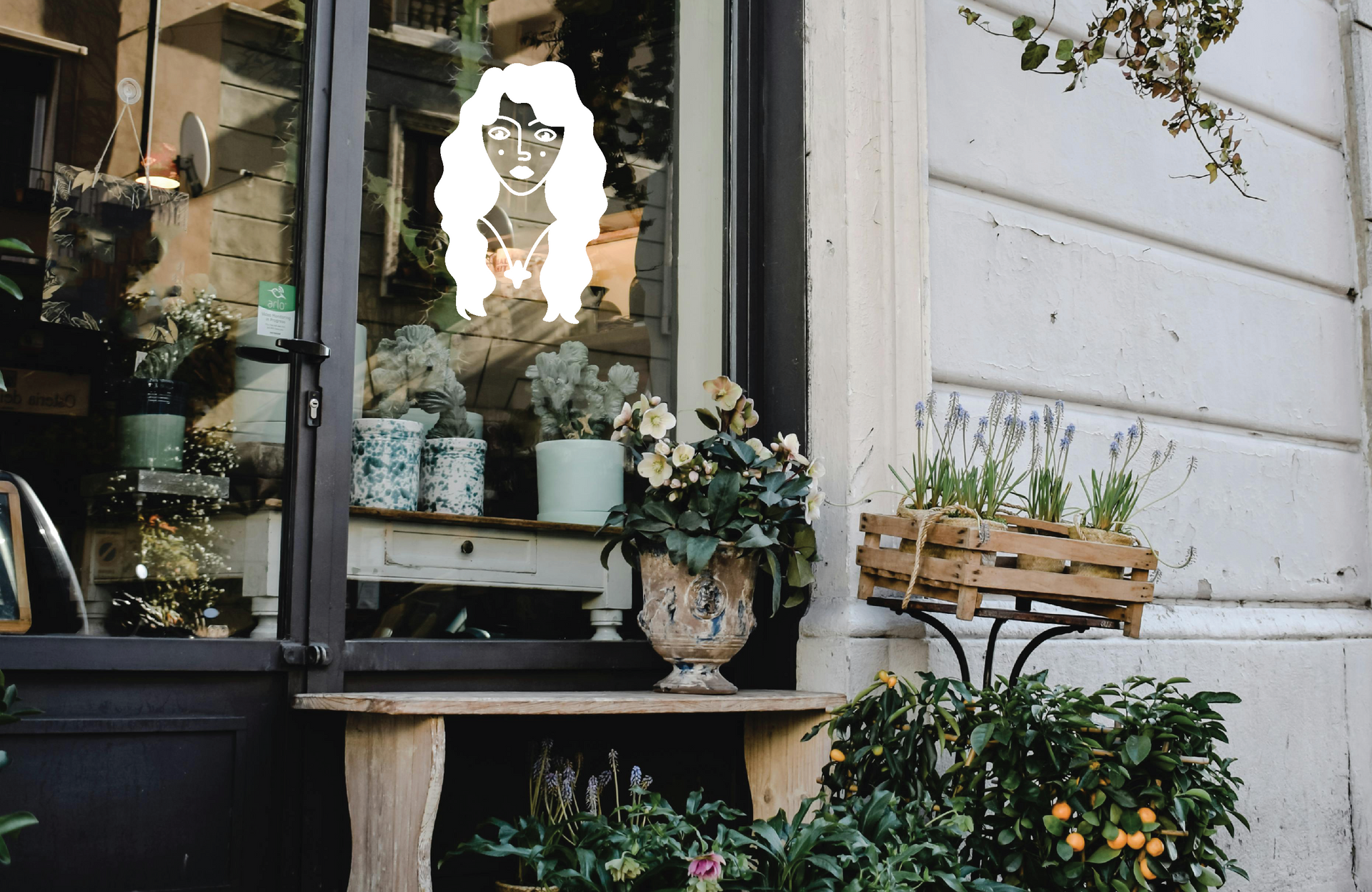 Exterior of an apothecary shop with plants, flowers, and a window sticker of House of Wayward Women's logo.