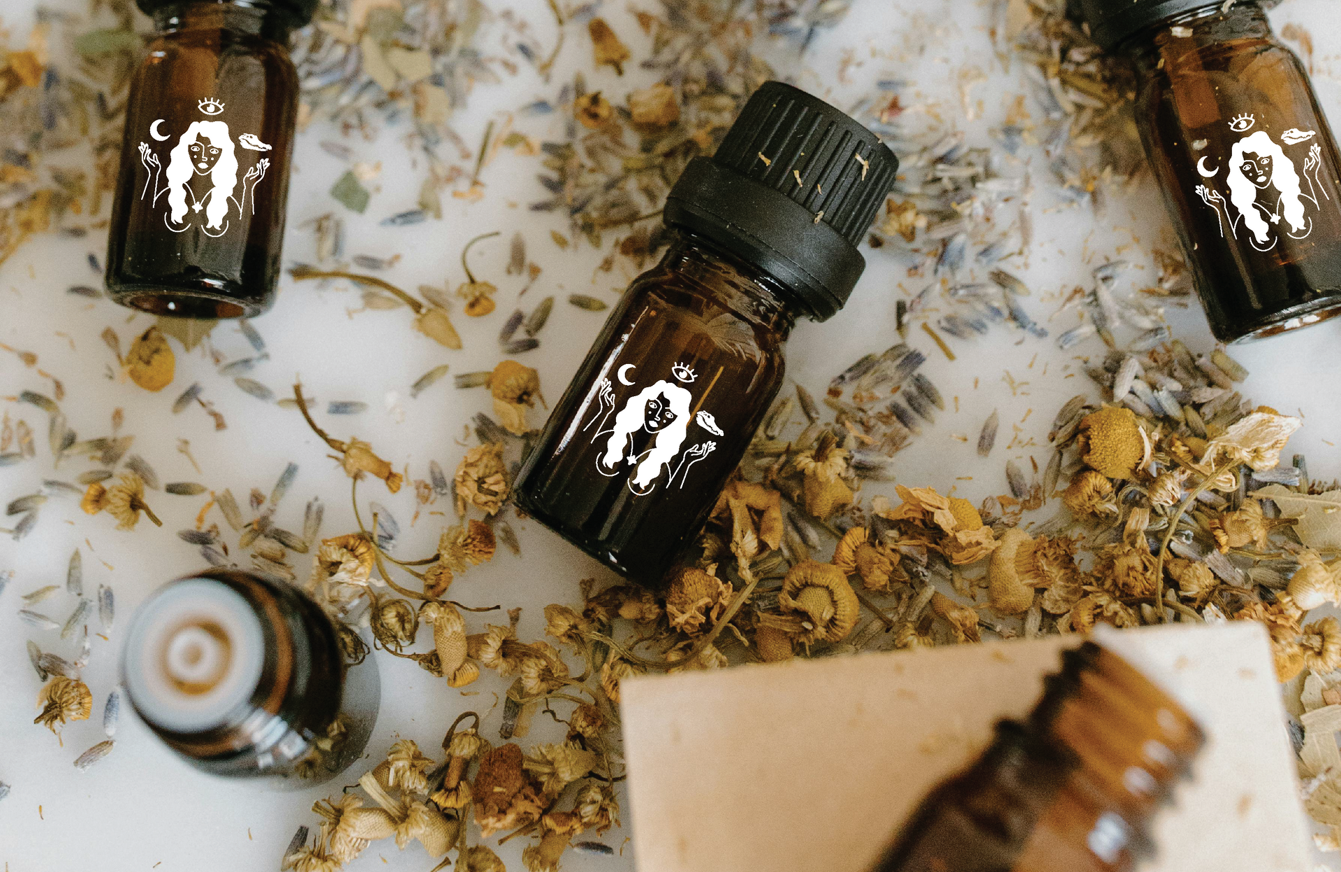 Small House of Wayward Women amber bottles of essential oil scattered amidst dried flowers on a white surface.