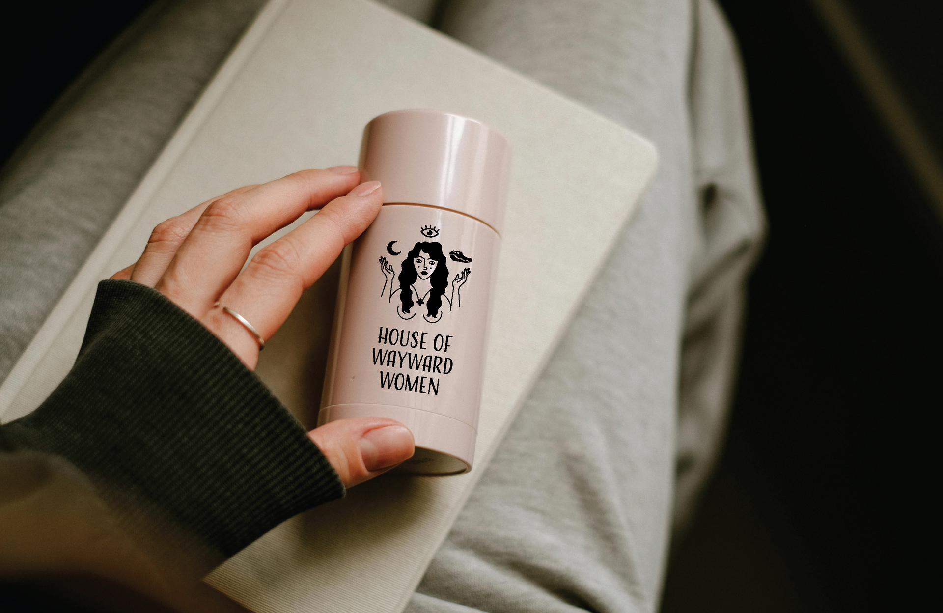 Person's hand holding a pink deodorant with a logo for House of Wayward Women with black illustration and text.