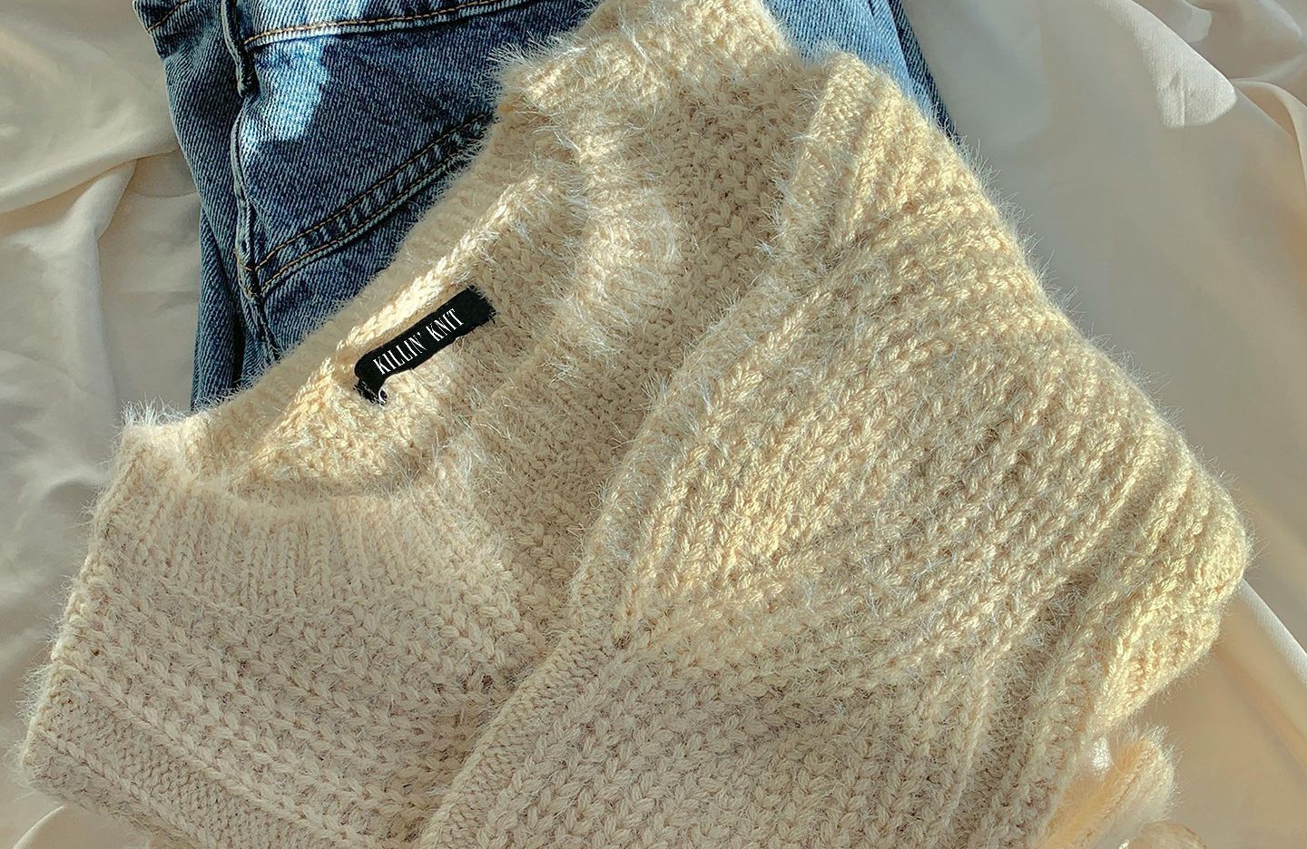 Cream-colored Killin' Knit knitted sweater and blue jeans on a white fabric background.