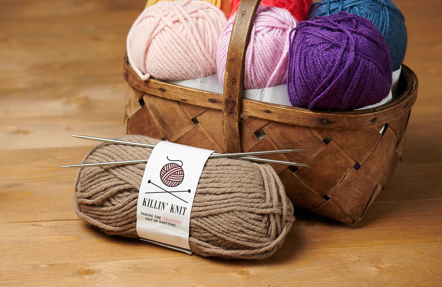 Basket of colorful Killin' Knit yarn balls with brown yarn and knitting needles in front.