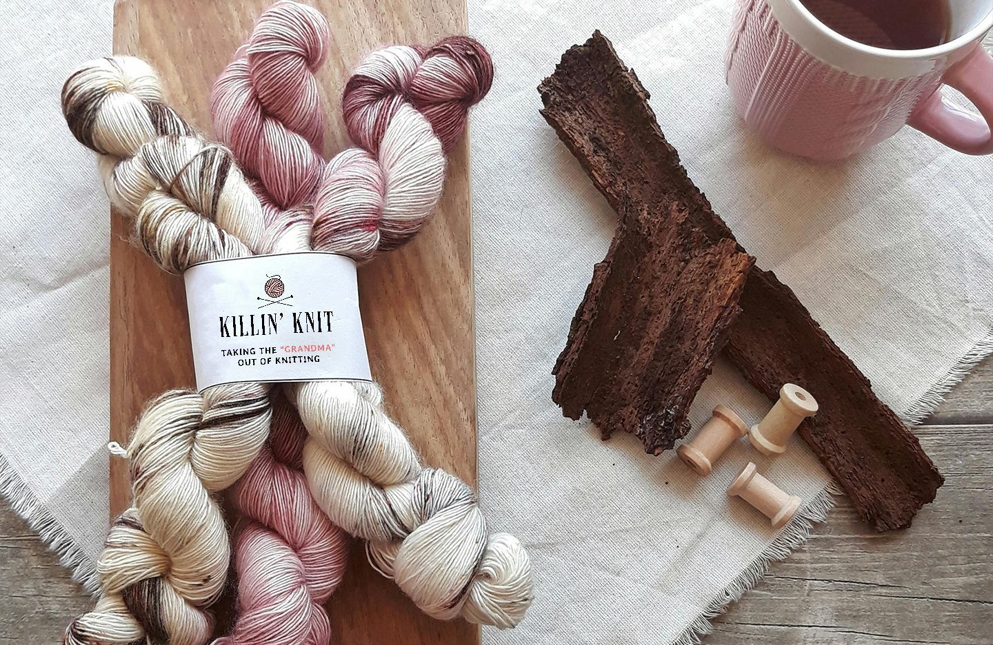 Killin' Knit skeins of hand-dyed yarn with a label, two pieces of dark bark, wooden spools, and a mug of tea.