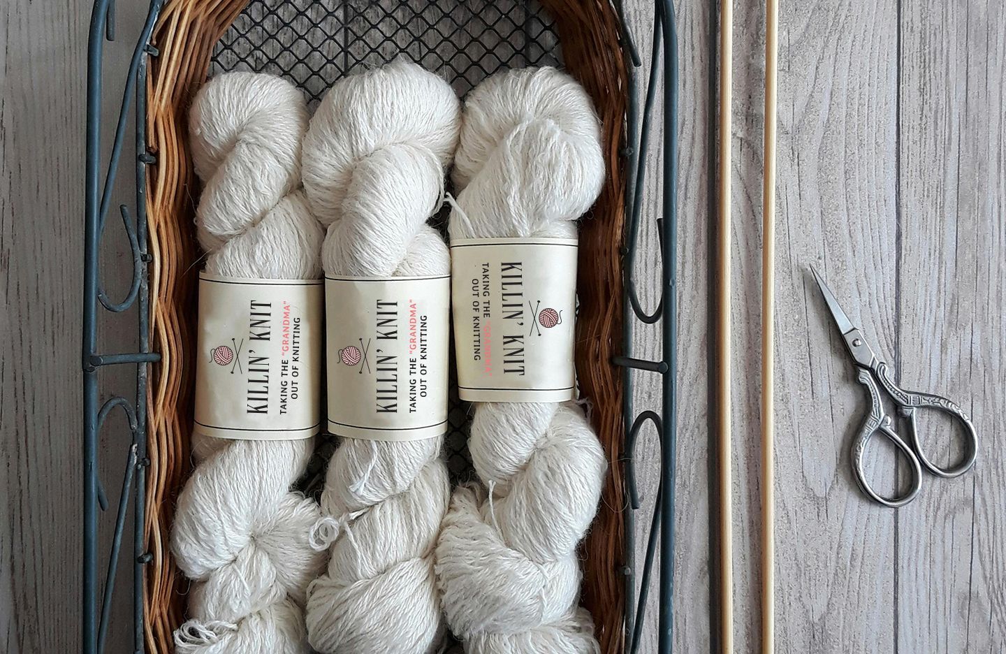 White Killin' Knit yarn skeins in a basket with knitting needles and scissors on a wooden surface.