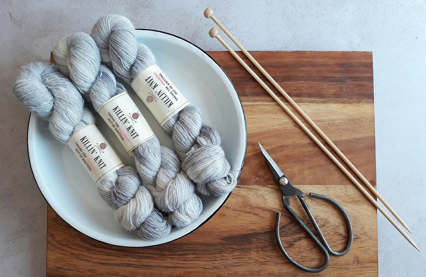 Skeins of gray Killin' Knit yarn in a white dish with knitting needles and scissors on a wooden board.