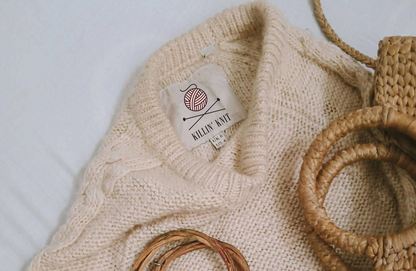 Cream-colored knit sweater with Killin' Knit label, near a woven bag and coiled rope, on a white surface.