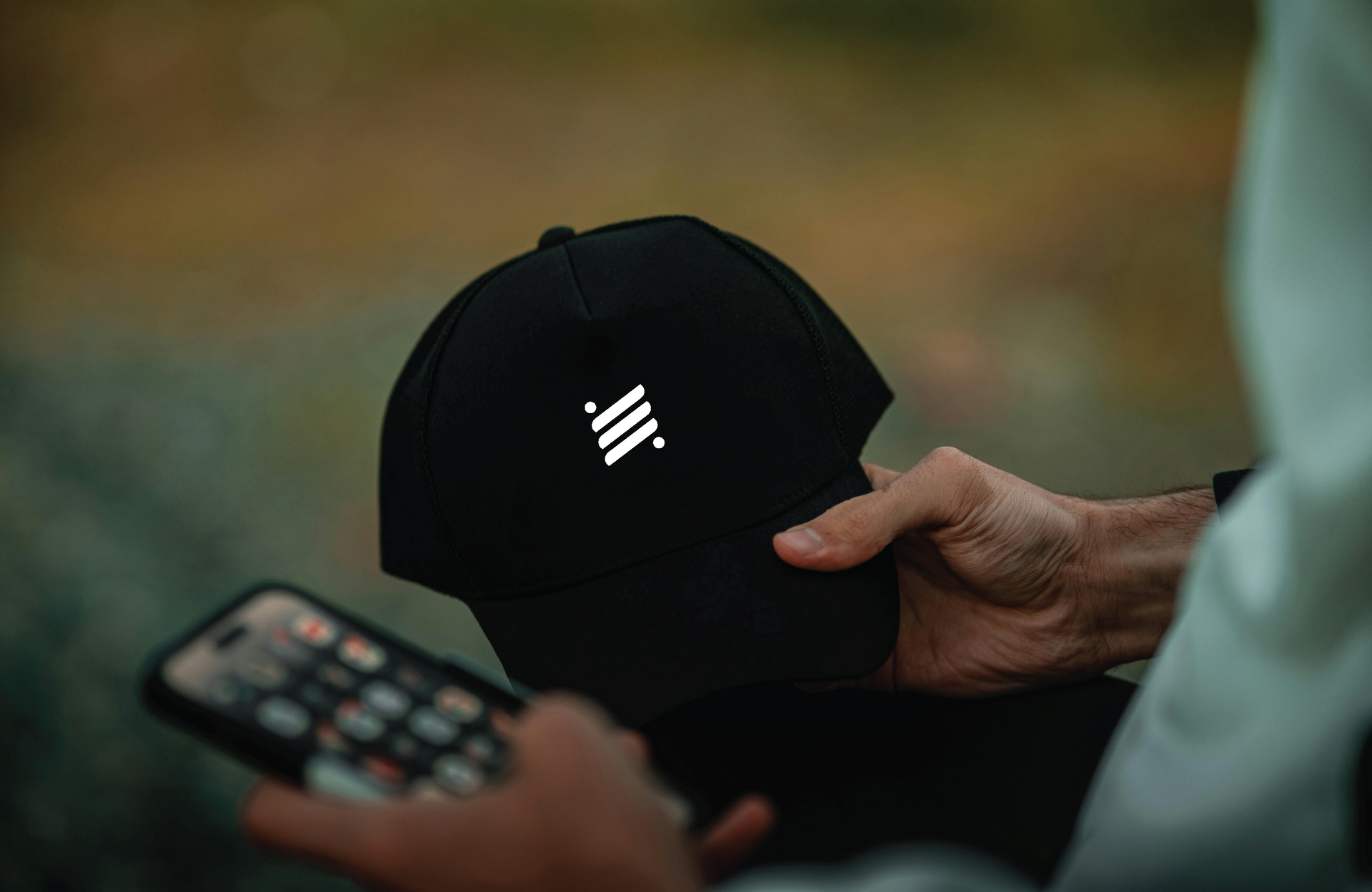 Person holding a black cap with a white Scout logo, with a blurred smartphone in view.