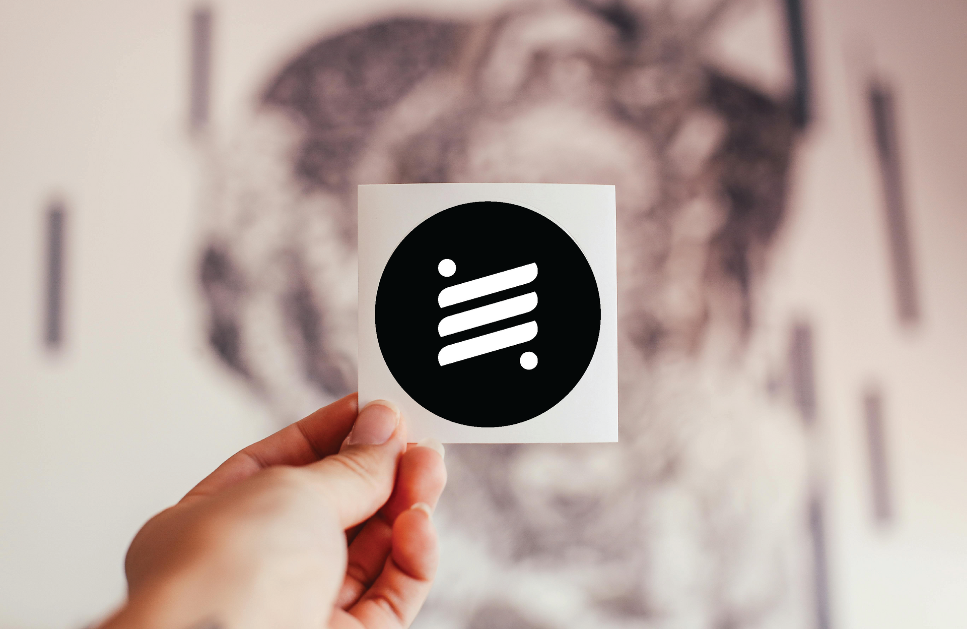 Hand holding a white sticker with a black circle and striped design in front of a blurry background.