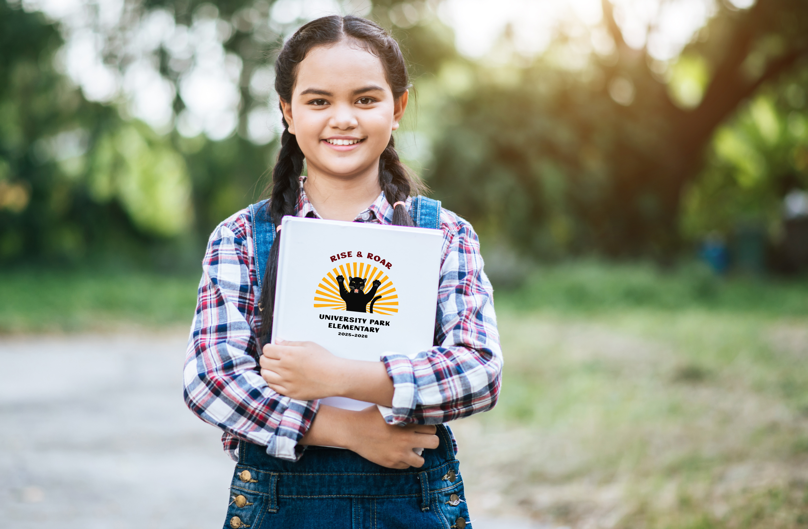 Girl with braids smiling, holding a book with a logo outdoors in sunlight.