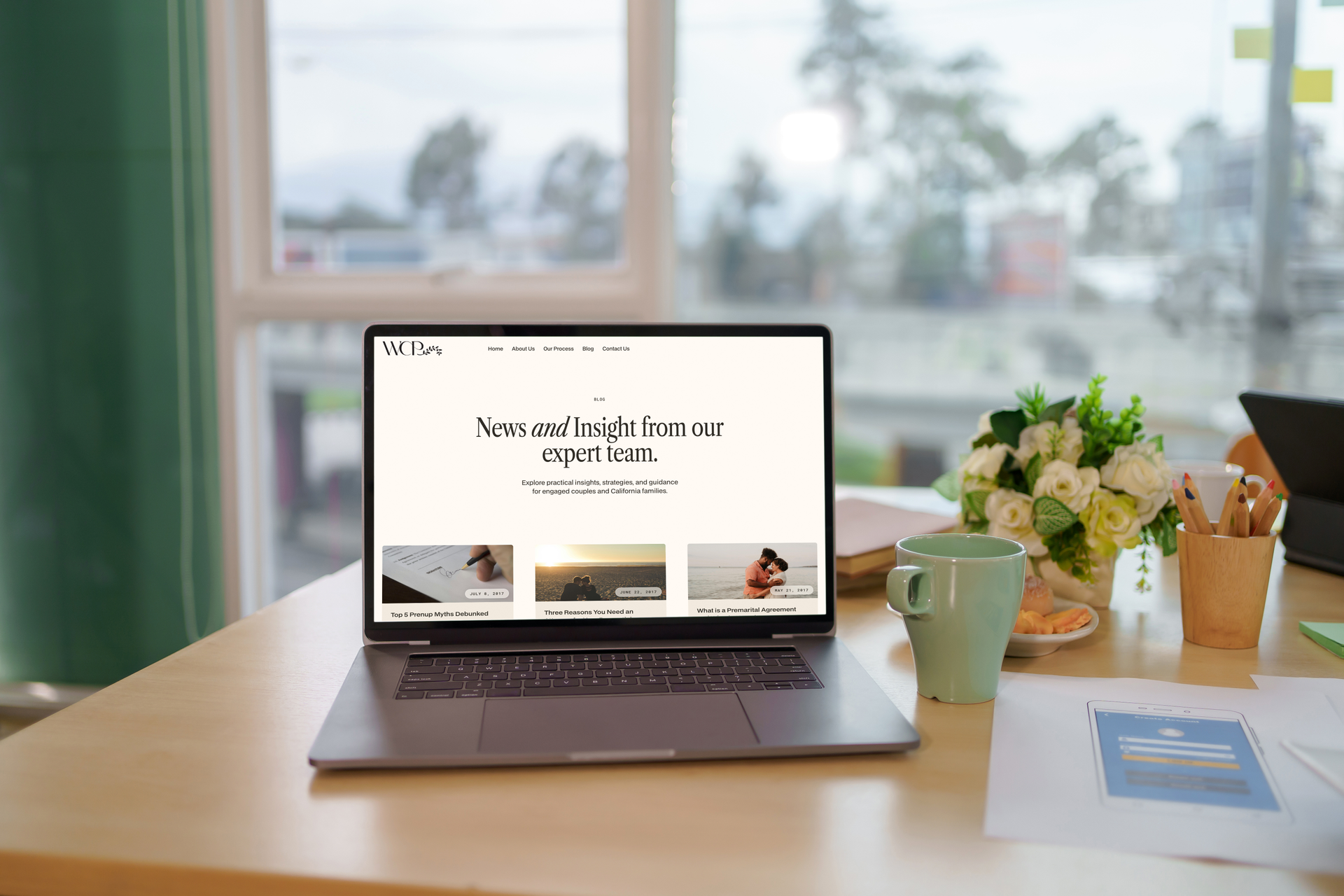 Laptop displaying a website on a desk, with a mug, flowers, and documents in an office setting.